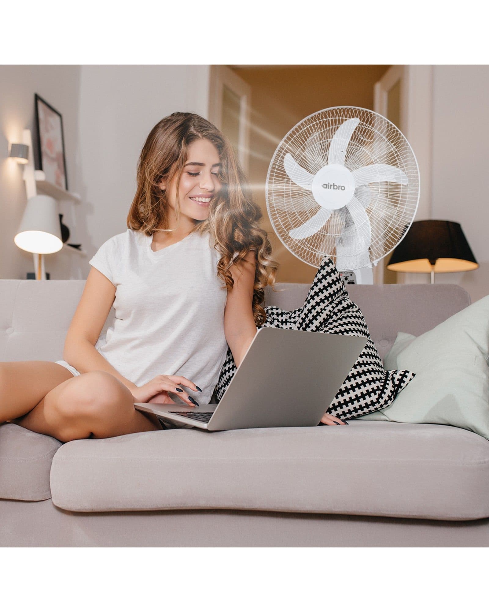 A smiling woman sits on a couch using a laptop while an Airbro FSA-180 pedestal fan blows air nearby.