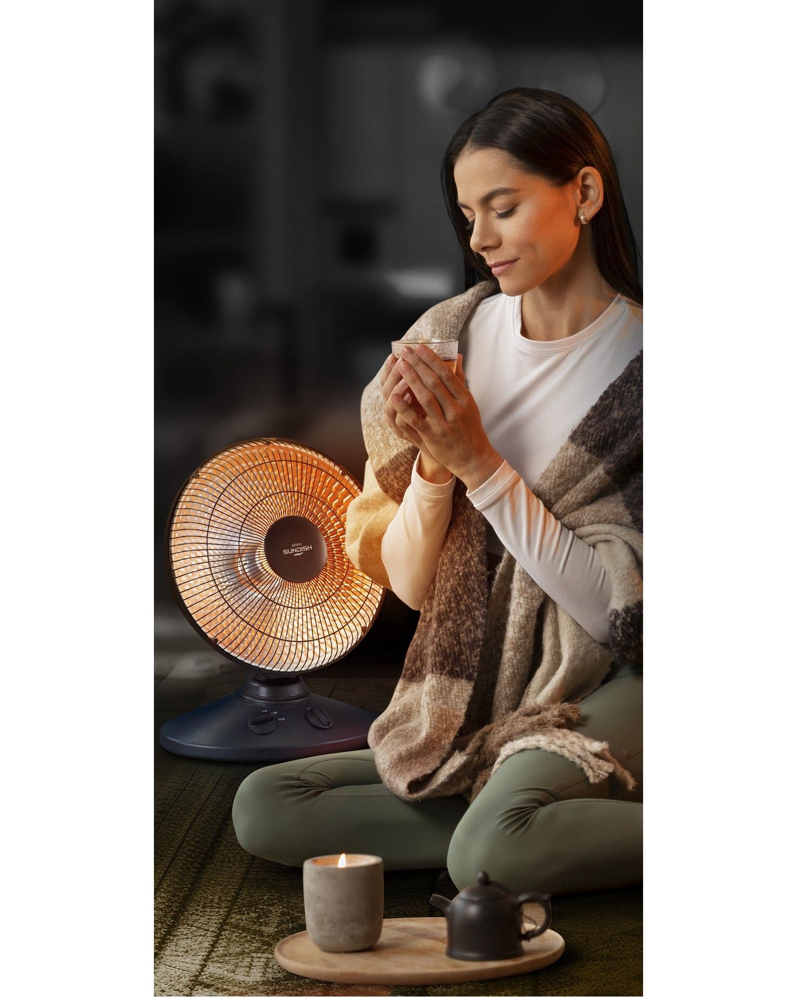 A woman wrapped in a blanket sits on the floor next to an Airbro HP-1000 heater, holding a warm drink.