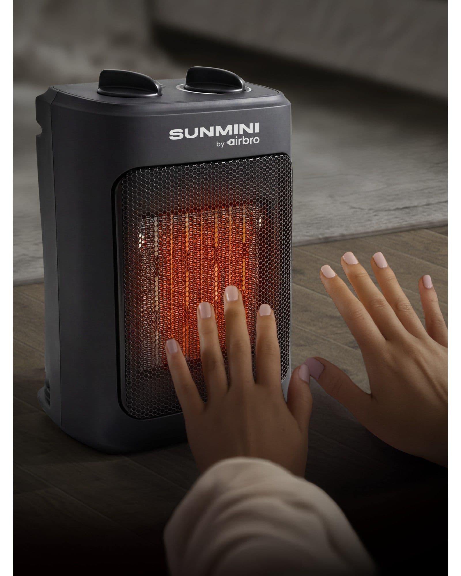 A person warming their hands in front of a glowing Airbro Sunmini portable space heater on a wooden floor.