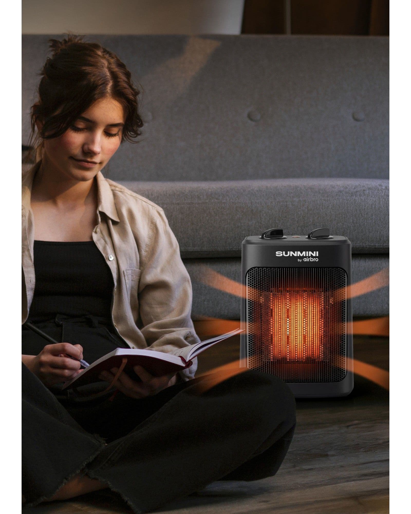 A woman sitting on the floor writing in a notebook next to an Airbro Sunmini space heater glowing with warmth.