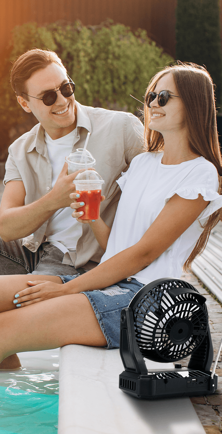 A young couple wearing sunglasses sits by a pool holding drinks next to a black Airbro RXF-40 portable desk fan.